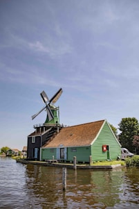 A traditional Dutch windmill with a green wooden building and red-tiled roof is situated next to a body of water. The sky is clear with a few clouds, and there are trees and additional structures in the background.