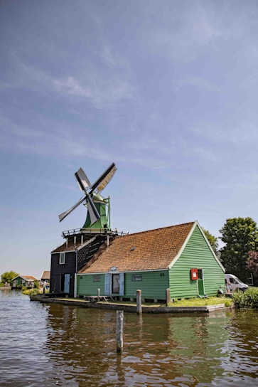 A traditional Dutch windmill with a green wooden building and red-tiled roof is situated next to a body of water. The sky is clear with a few clouds, and there are trees and additional structures in the background.