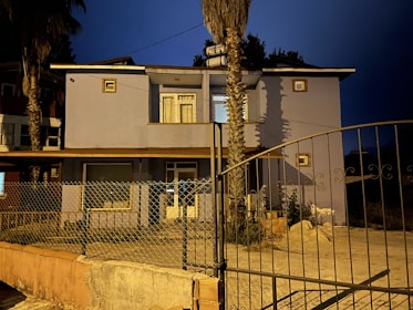 Two-story house with blue exterior walls, seen at night under artificial lighting. The house features small square windows, a larger window below a balcony, and a gated entrance with a mesh fence enclosing a small yard. Two tall palm trees are positioned in front of the house, casting shadows on its facade.