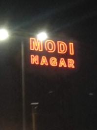 The image features a large, illuminated sign with the words 'MODI NAGAR' displayed in bright red neon lights against a dark background. The sign is positioned on a tall pole with additional light sources visible behind it, casting a glow around the letters.