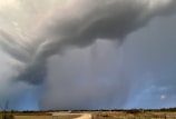 A swirling storm cloud dominates the sky, creating a dramatic and imposing atmosphere. The landscape below is flat, with a stretch of road and some vegetation visible. The storm appears to be intense, with dark and heavy clouds suggesting possible rainfall.