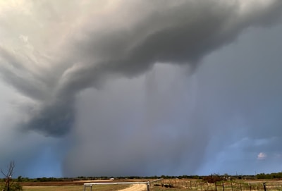 A swirling storm cloud dominates the sky, creating a dramatic and imposing atmosphere. The landscape below is flat, with a stretch of road and some vegetation visible. The storm appears to be intense, with dark and heavy clouds suggesting possible rainfall.
