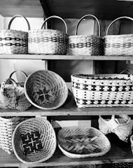 A collection of woven baskets displayed on wooden shelves. Each basket features intricate patterns and is crafted in a traditional style. The baskets vary in shape and size, ranging from round to rectangular, and are made from natural materials.