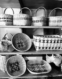 A collection of woven baskets displayed on wooden shelves. Each basket features intricate patterns and is crafted in a traditional style. The baskets vary in shape and size, ranging from round to rectangular, and are made from natural materials.