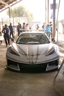 A sleek, customized sports car with 'Liberty Walk' branding is prominently displayed in a sunny outdoor setting. A few people are gathered around, some in casual attire and others appearing to socialize or admire the vehicle. The environment seems like a car meet or auto show.