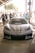 A sleek, customized sports car with 'Liberty Walk' branding is prominently displayed in a sunny outdoor setting. A few people are gathered around, some in casual attire and others appearing to socialize or admire the vehicle. The environment seems like a car meet or auto show.