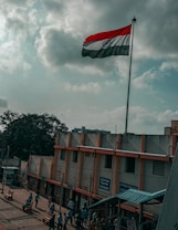 A tall flagpole with an Indian flag flies prominently against a cloudy sky. Below, there is a building with signs in multiple languages, and several people walk along a pathway beside it. A large tree is visible in the background, and there are shadows cast by the structures and people.