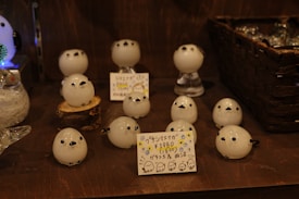 A display of small, spherical, white figurines with black dots for eyes and feet, arranged on a wooden shelf. Some figurines are placed on small stands or tree stumps, and there are handwritten price tags with Japanese text in front of them.