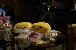 A table with various items including plush toys and packaged goods. Two yellow plushies with simple facial expressions are in clear plastic bags. Beside them, baskets hold various items, including wrapped snacks and packages featuring colorful designs. A plush toy resembling a mouse character is also visible on the left.