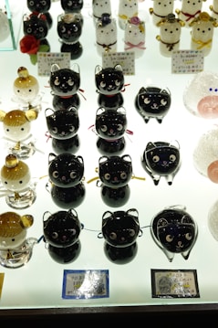 An assortment of small, glossy figurines resembling cats and other animals, displayed on a brightly lit surface. The figures have various colors, including black, white, and golden, and feature playful expressions. Some have decorative elements like bows and flowers.