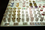 Glass figures of various animals are neatly arranged on a well-lit display table. These figures include shapes resembling bears, dogs, and pigs, each with distinct facial expressions and colors. The figures are in organized rows, and some have additional decorations or colored elements. Labels with text are placed in front of some of the figures, possibly indicating their price or name.