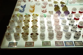 Glass figures of various animals are neatly arranged on a well-lit display table. These figures include shapes resembling bears, dogs, and pigs, each with distinct facial expressions and colors. The figures are in organized rows, and some have additional decorations or colored elements. Labels with text are placed in front of some of the figures, possibly indicating their price or name.