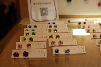 A glass display case contains an organized arrangement of small, colorful stones or earrings placed on cards. The interior lighting highlights the glossy and vibrant nature of the items, which come in various colors such as blue, green, yellow, and pink. The cards are printed with text and are neatly arranged in rows, showcasing the items effectively.
