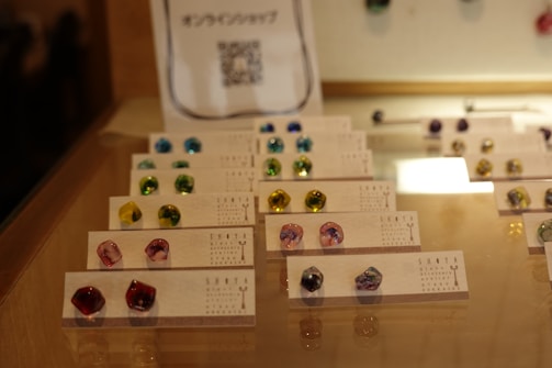 A glass display case contains an organized arrangement of small, colorful stones or earrings placed on cards. The interior lighting highlights the glossy and vibrant nature of the items, which come in various colors such as blue, green, yellow, and pink. The cards are printed with text and are neatly arranged in rows, showcasing the items effectively.