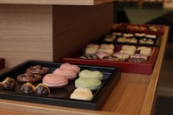 A tray of assorted pastries is displayed on wooden shelves. There are macarons in pink and green on one tray, while another tray contains pretzel-shaped chocolates. In the background, a variety of pastries with icing and chocolate drizzle are arranged on another tray.
