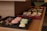 A tray of assorted pastries is displayed on wooden shelves. There are macarons in pink and green on one tray, while another tray contains pretzel-shaped chocolates. In the background, a variety of pastries with icing and chocolate drizzle are arranged on another tray.