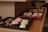 A tray of assorted pastries is displayed on wooden shelves. There are macarons in pink and green on one tray, while another tray contains pretzel-shaped chocolates. In the background, a variety of pastries with icing and chocolate drizzle are arranged on another tray.