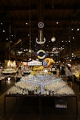 A well-lit indoor market or store features a central display of various glass bottles and ornaments arranged on a table. Above the table, a decorative anchor and a life ring are suspended from the ceiling. The ambiance is warm with wooden beams and numerous hanging lights. Several people are browsing the items, adding a bustling yet cozy atmosphere to the setting.