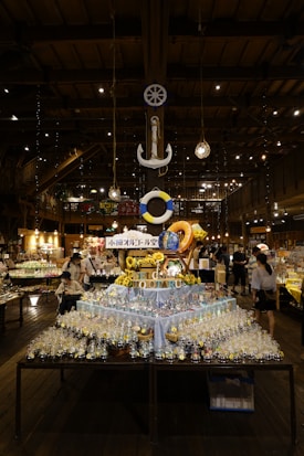 A well-lit indoor market or store features a central display of various glass bottles and ornaments arranged on a table. Above the table, a decorative anchor and a life ring are suspended from the ceiling. The ambiance is warm with wooden beams and numerous hanging lights. Several people are browsing the items, adding a bustling yet cozy atmosphere to the setting.