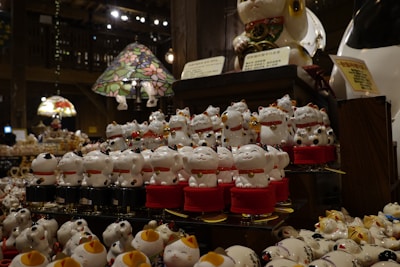 A variety of ceramic lucky cat figurines are displayed on shelves in a store. They are mostly white, adorned with red collars and gold details. Some are standing while others are seated, with paws raised in traditional gestures. The shelves are under warm ambient lighting, and there are colorful stained glass lamps visible in the background.