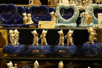 A collection of ceramic cat figurines playing musical instruments is displayed on shelves. The figures are white with blue bases and are adorned with blue ribbons. Each figure holds a golden instrument, including trumpets and saxophones. Heart-shaped frames with musical notes are placed behind the figurines, adding to the musical theme.