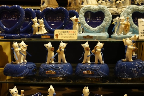 A collection of ceramic cat figurines playing musical instruments is displayed on shelves. The figures are white with blue bases and are adorned with blue ribbons. Each figure holds a golden instrument, including trumpets and saxophones. Heart-shaped frames with musical notes are placed behind the figurines, adding to the musical theme.