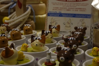 Several small, cute figurines are arranged in white cups. The figurines consist of animals like brown bears and cats with stripes on their heads, and small yellow ducks. The background shows ropes, wooden objects, and possibly a sign with illustrations.