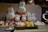 A display of various plush toys resembling a white bunny with a simplistic facial expression and orange clothing. The plush toys are arranged in multiple sizes, sitting in baskets on a table with a cloth draped over it. A person wearing a mask is visible on the right side, standing behind the display.