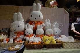 A display of various plush toys resembling a white bunny with a simplistic facial expression and orange clothing. The plush toys are arranged in multiple sizes, sitting in baskets on a table with a cloth draped over it. A person wearing a mask is visible on the right side, standing behind the display.