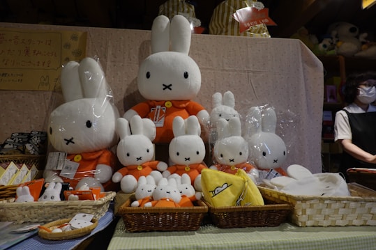 A display of various plush toys resembling a white bunny with a simplistic facial expression and orange clothing. The plush toys are arranged in multiple sizes, sitting in baskets on a table with a cloth draped over it. A person wearing a mask is visible on the right side, standing behind the display.