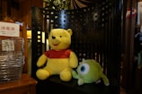 Two plush toys are placed on a large, black, lattice-backed chair. One toy resembles a bear in a red shirt, while the other is a green character with a single large eye. A stack of wire baskets and a sign with Japanese text are visible on the left. The setting includes warm, wooden tones with a plant in the background.