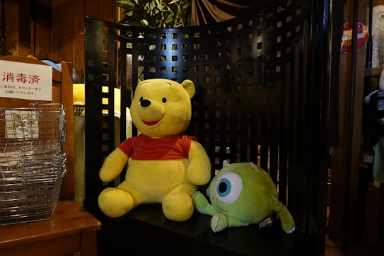 Two plush toys are placed on a large, black, lattice-backed chair. One toy resembles a bear in a red shirt, while the other is a green character with a single large eye. A stack of wire baskets and a sign with Japanese text are visible on the left. The setting includes warm, wooden tones with a plant in the background.