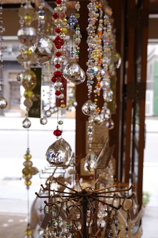 Sparkling crystal and glass ornaments of various colors, including red, blue, and yellow, hang from the ceiling. These intricate decorations glisten in the natural light, showcasing their multifaceted surfaces. They are displayed in a shop setting, with some metal stands holding keychains and accessories in the foreground.