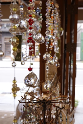 Sparkling crystal and glass ornaments of various colors, including red, blue, and yellow, hang from the ceiling. These intricate decorations glisten in the natural light, showcasing their multifaceted surfaces. They are displayed in a shop setting, with some metal stands holding keychains and accessories in the foreground.