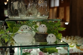 Glass figurines resembling birds are placed on a transparent shelf, surrounded by leafy decorations. One of the figurines rests on a small wooden base, while others have bright red cheeks and small black eyes. The background shows a warm, softly lit indoor setting with more glass items displayed.
