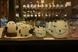 A collection of decorative glass figures resembling animals, including round, white shapes with black accents, arranged on a shelf. The background consists of an assortment of glass objects, adding a colorful and artistic ambiance to the scene.
