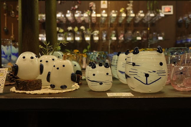 A collection of decorative glass figures resembling animals, including round, white shapes with black accents, arranged on a shelf. The background consists of an assortment of glass objects, adding a colorful and artistic ambiance to the scene.
