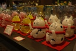 A display of decorative cat figurines, each crafted from glass and featuring a white and black spotted design. The cats have red collars with gold bells and are standing on red mats. In the background, there are more glass items, including similar cat figurines in pink and yellow. The image depicts a variety of glassware, likely in a store setting.