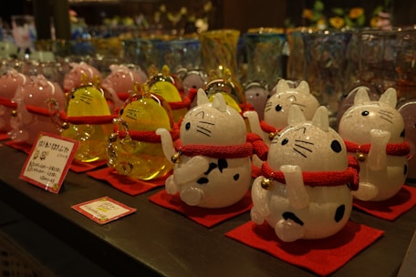 A display of decorative cat figurines, each crafted from glass and featuring a white and black spotted design. The cats have red collars with gold bells and are standing on red mats. In the background, there are more glass items, including similar cat figurines in pink and yellow. The image depicts a variety of glassware, likely in a store setting.