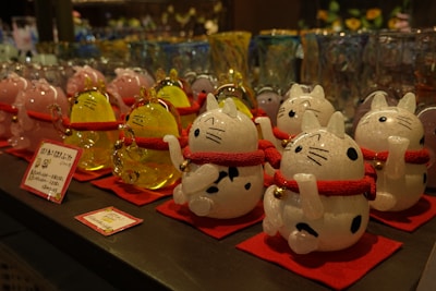 A display of decorative cat figurines, each crafted from glass and featuring a white and black spotted design. The cats have red collars with gold bells and are standing on red mats. In the background, there are more glass items, including similar cat figurines in pink and yellow. The image depicts a variety of glassware, likely in a store setting.