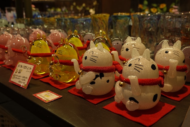 A display of decorative cat figurines, each crafted from glass and featuring a white and black spotted design. The cats have red collars with gold bells and are standing on red mats. In the background, there are more glass items, including similar cat figurines in pink and yellow. The image depicts a variety of glassware, likely in a store setting.