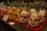 A display of decorative cat figurines, each crafted from glass and featuring a white and black spotted design. The cats have red collars with gold bells and are standing on red mats. In the background, there are more glass items, including similar cat figurines in pink and yellow. The image depicts a variety of glassware, likely in a store setting.