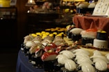 A display of various types of sushi is arranged on a table. The sushi pieces are placed on small pedestals, showcasing a variety of fish and seafood toppings including octopus, tuna, and shrimp. The environment appears to be dimly lit, suggesting an elegant or upscale setting. A sign with Japanese writing is visible, adding an authentic touch to the display.