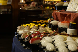 A display of various types of sushi is arranged on a table. The sushi pieces are placed on small pedestals, showcasing a variety of fish and seafood toppings including octopus, tuna, and shrimp. The environment appears to be dimly lit, suggesting an elegant or upscale setting. A sign with Japanese writing is visible, adding an authentic touch to the display.