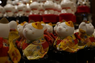 A collection of ceramic or porcelain lucky cat figurines wearing colorful, patterned kimonos. The cats have a cheerful expression, with one paw raised. The background shows more of these figurines, creating a crowded and lively scene.