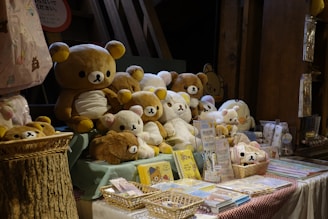 A collection of various stuffed bear toys displayed on a table, surrounded by related merchandise and stationery items. The setting has a cozy and warm atmosphere, with natural wooden elements adding to the charm.