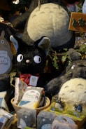 A variety of plush toys and merchandise featuring characters from a popular animated movie are displayed. The toys include large and small plush figures of a large grey creature with a white belly and a smaller black cat with a red bow. Surrounding these are various packaged items with character designs.