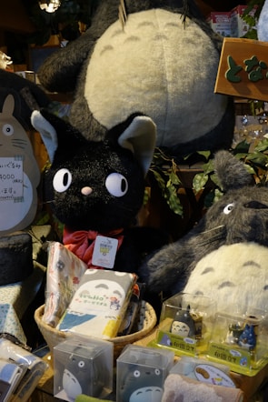 A variety of plush toys and merchandise featuring characters from a popular animated movie are displayed. The toys include large and small plush figures of a large grey creature with a white belly and a smaller black cat with a red bow. Surrounding these are various packaged items with character designs.