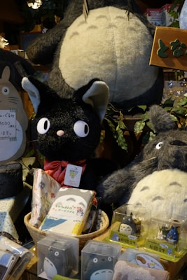 A variety of plush toys and merchandise featuring characters from a popular animated movie are displayed. The toys include large and small plush figures of a large grey creature with a white belly and a smaller black cat with a red bow. Surrounding these are various packaged items with character designs.