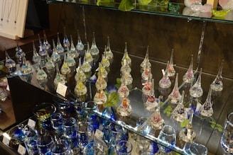 Variety of delicate glass ornaments with long, slender necks displayed on glass shelves. The ornaments are filled with colorful materials like beads or glitter, creating a shimmering effect under the light. Several leaves and other decorative glass items such as bowls and marbles are also arranged on the shelves.